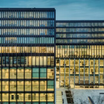 Verlicht kantoorgebouw met veel ramen, als beeld voor financiële macht, belastingroutes en besluiten achter gesloten deuren.