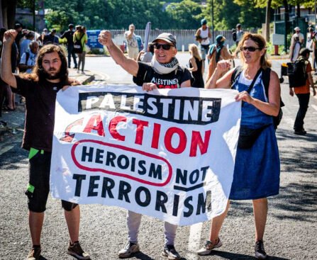 Drie demonstranten houden een spandoek vast met de tekst “Palestine Action, heroism not terrorism” tijdens een protest op straat.