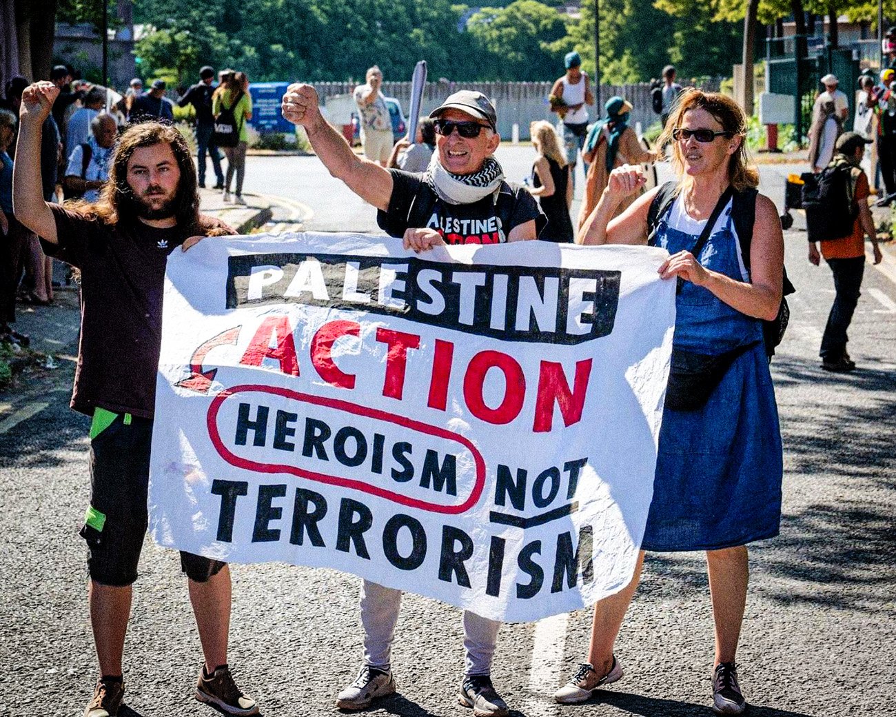 Drie demonstranten houden een spandoek vast met de tekst “Palestine Action, heroism not terrorism” tijdens een protest op straat.