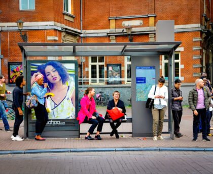 Mensen wachten bij een tramhalte op het Leidseplein in Amsterdam, naast een grote reclameposter en een kaart van het openbaar vervoer.