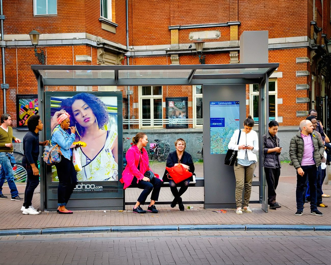 Mensen wachten bij een tramhalte op het Leidseplein in Amsterdam, naast een grote reclameposter en een kaart van het openbaar vervoer.